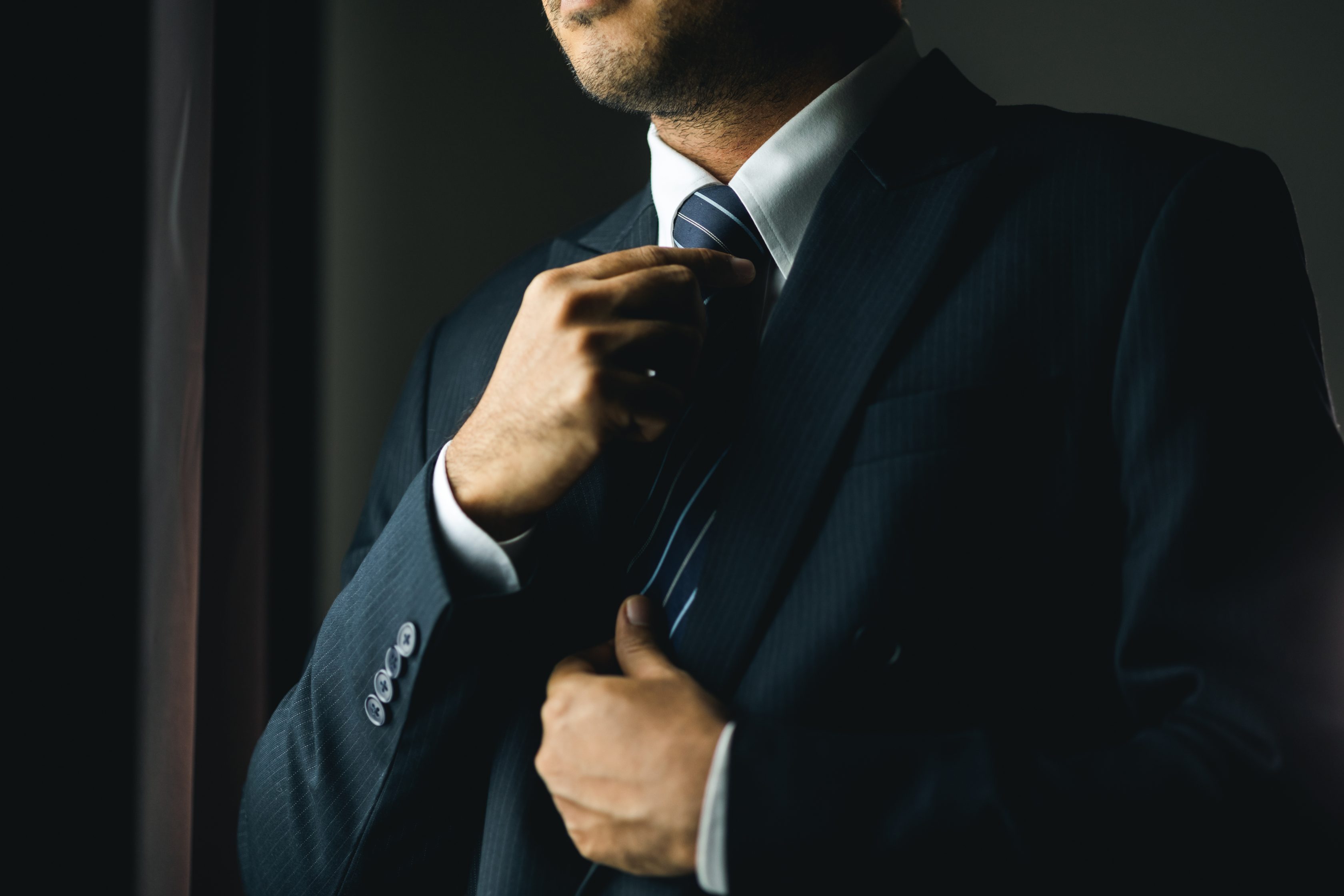 Confident businessman buttoning or adjust classic blue suit near window in hotel room at the morning. Handsome man wearing a nice suit on wedding day.